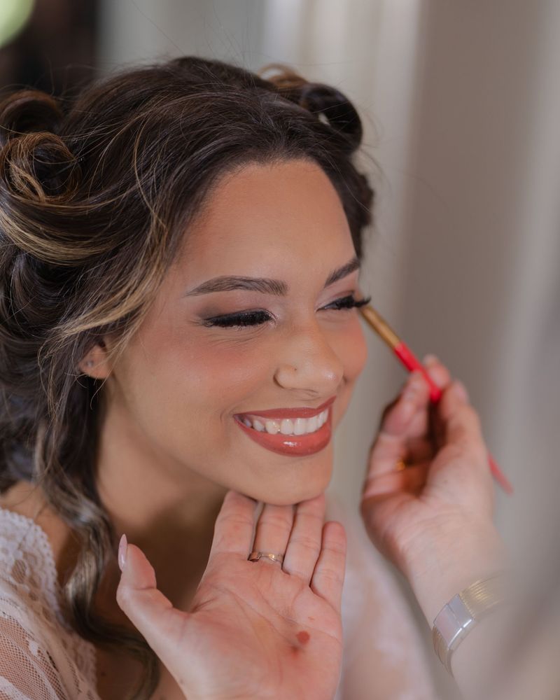 Smiling bride in a lace robe having her eye makeup finished with a brush, her hair curled and pinned up