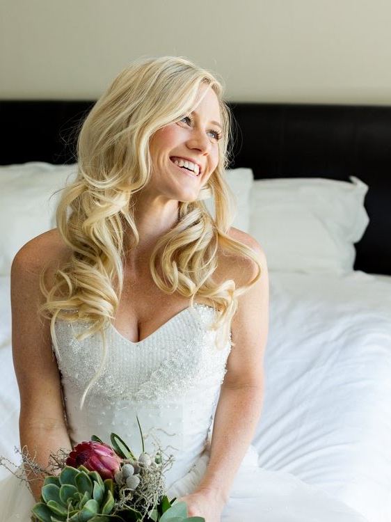 Laughing bride with long blonde Hollywood waves, sitting on a bed with a protea bouquet