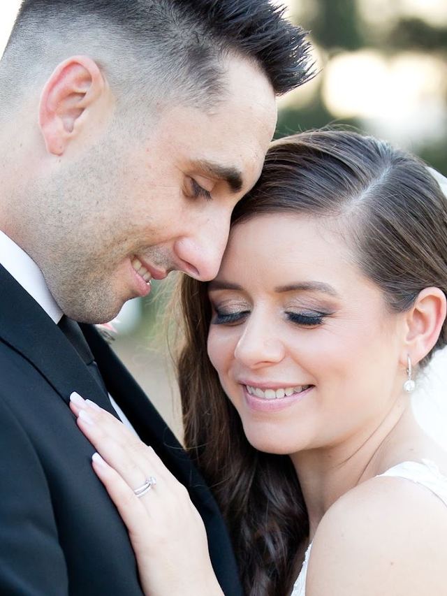Groom resting his head on his bride, who has a sleek side-swept style with a veil and soft glam makeup