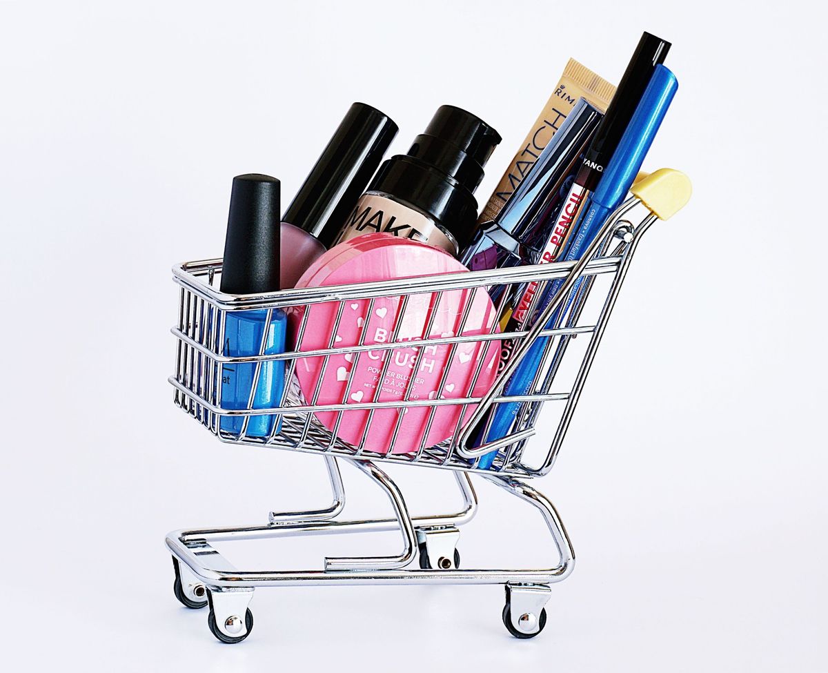 Miniature shopping trolley filled with makeup, including a pink blush compact, foundation, mascara, nail polish and eyeliner pencils