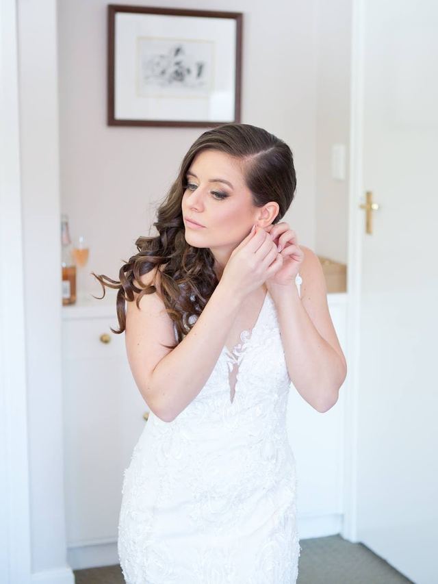 Bride putting on her earring, sleek side-swept hair in long loose curls and soft smoky eyes