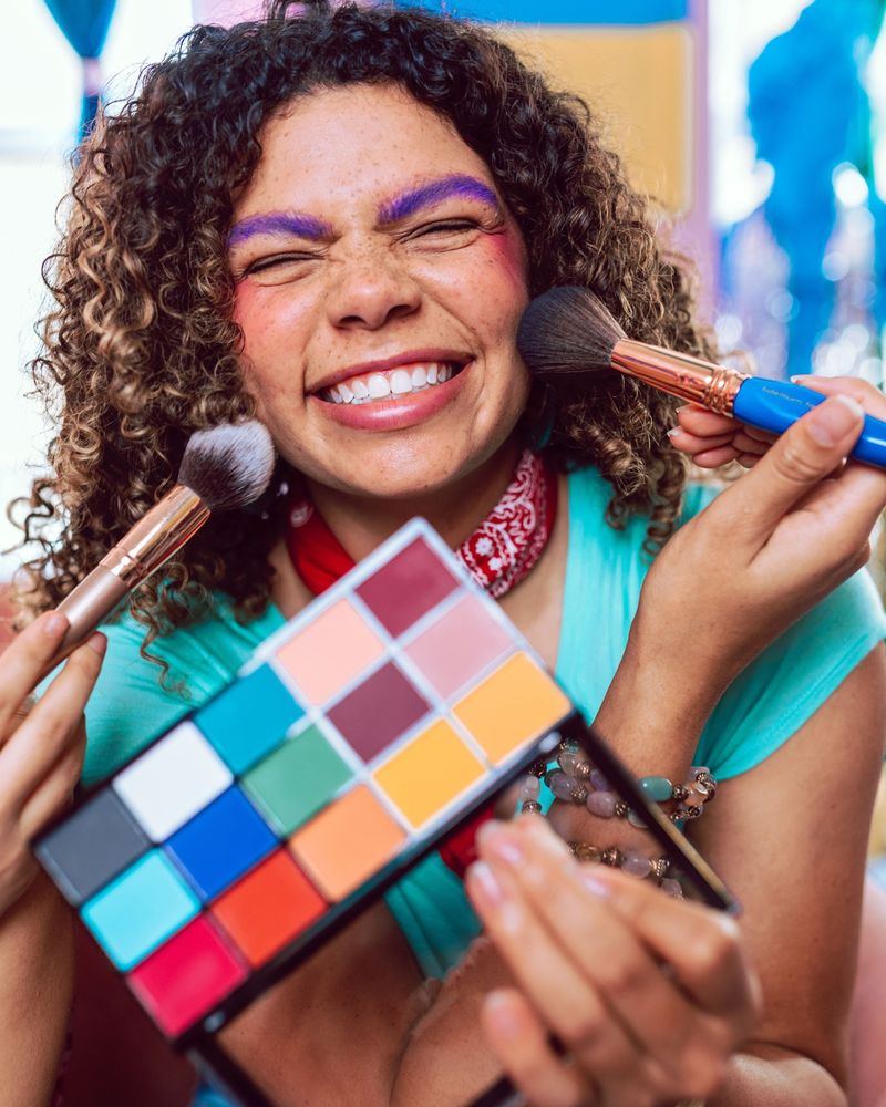 Laughing woman with curly hair and purple brows holding a colourful makeup palette, with two brushes at her cheeks