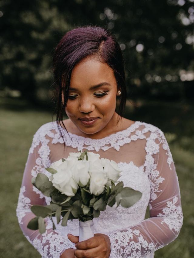 Bride in a long-sleeve lace gown looking down at her white rose bouquet, with violet-toned hair and gold eyeshadow