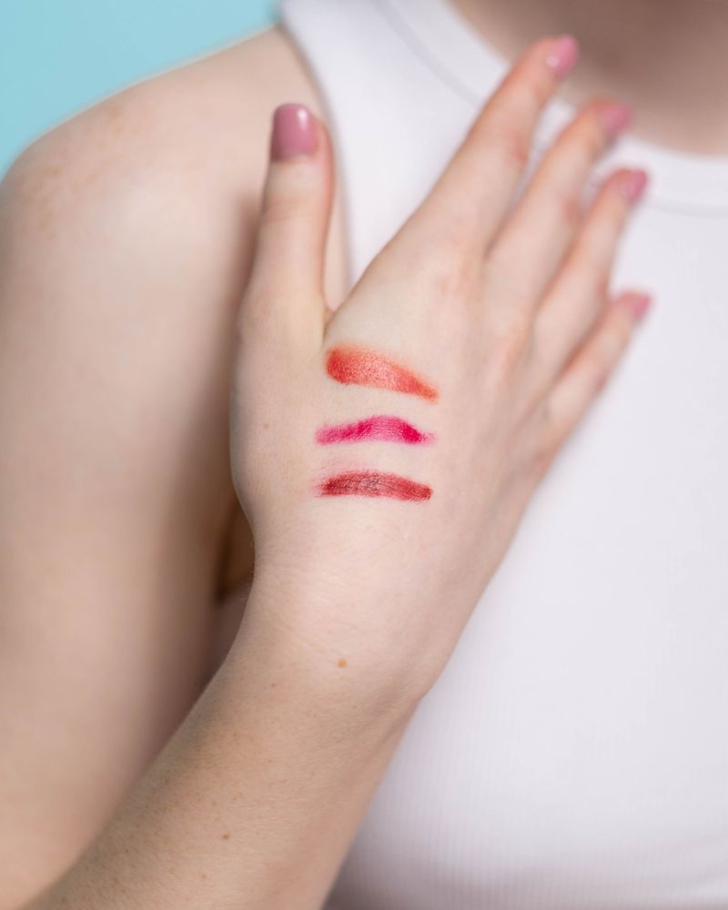 Three lip colour swatches in coral orange, hot pink and deep red on the back of a hand