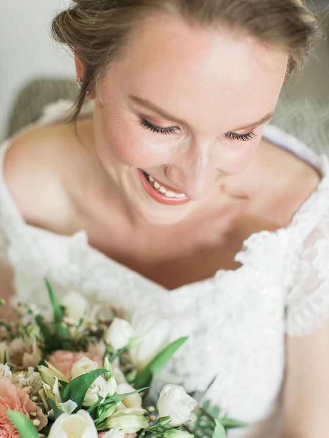Bride smiling down at her peach and white bouquet, soft natural makeup with a rosy glow