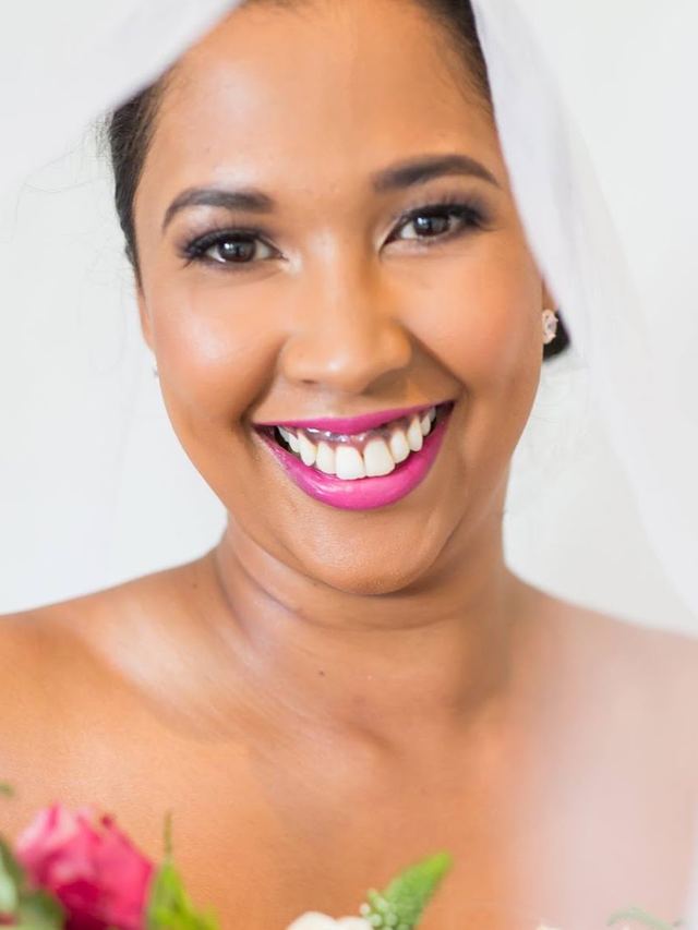 Smiling bride with bold pink lipstick under a sheer veil, holding a bouquet of pink roses