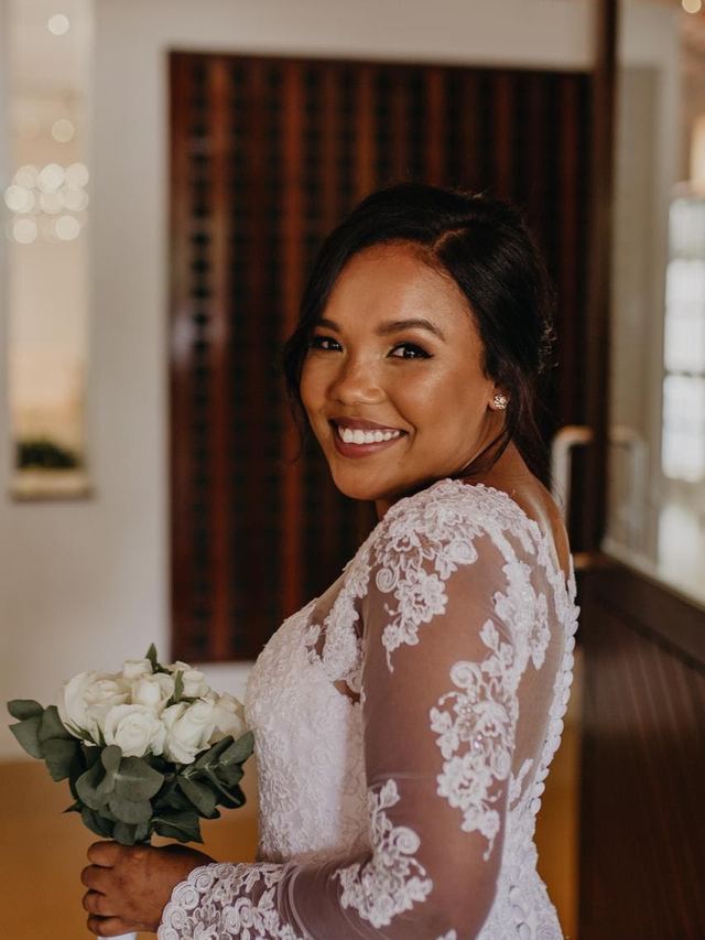 Bride in a lace long-sleeve gown smiling over her shoulder, glowing makeup and a sleek low updo